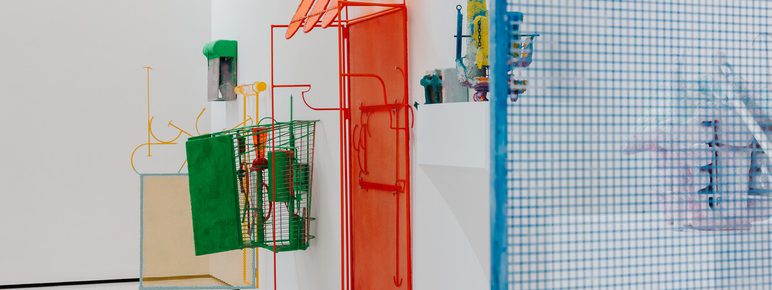 An art installation featuring colorful wire sculptures mounted on a white wall. The pieces include a red outline of a house, green and yellow structures, and various abstract forms, creating a playful and vibrant atmosphere in the gallery space.