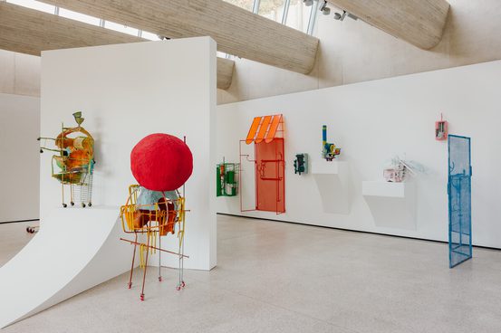 An art gallery featuring colorful sculptures made of metal and other materials. The pieces include abstract forms like a large red sphere, a house shape, and various whimsical structures, all displayed against white walls in a modern space.