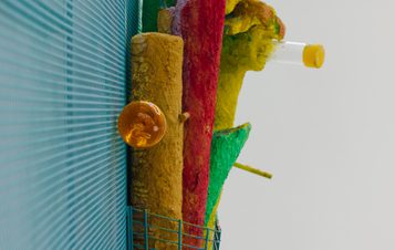 An abstract sculpture featuring vibrant colors like red, yellow, and green. It includes textured cylindrical forms and a transparent tube with a yellow liquid. The background is a blue grid pattern, adding depth to the composition.