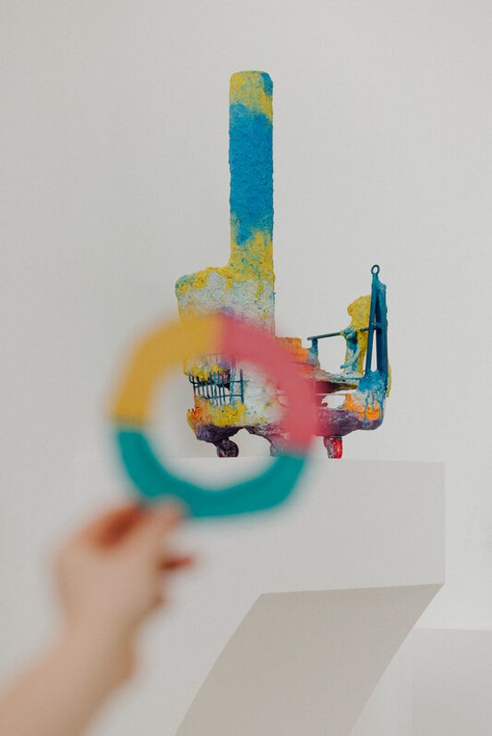 A colorful abstract sculpture stands on a white pedestal, featuring vibrant blue, yellow, and red hues. In the foreground, a hand holds a circular, multicolored frame, partially obscuring the sculpture, creating a playful visual effect.