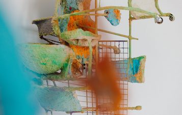 An abstract sculpture composed of textured, colorful materials in shades of green, orange, and blue. The piece features various shapes and forms, including bowls and grids, mounted on a wall, creating a dynamic visual composition.