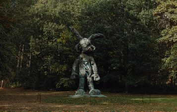 A large bronze sculpture of a humanoid rabbit stands in a wooded area. The figure has exaggerated features, including oversized ears and a playful stance, surrounded by lush green trees and grass, creating a whimsical atmosphere.