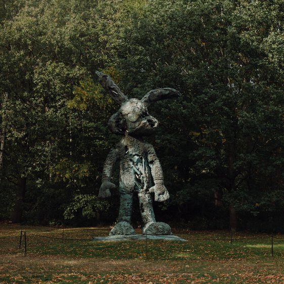 A large bronze sculpture of a humanoid rabbit stands in a wooded area. The figure has exaggerated features, including oversized ears and a playful stance, surrounded by lush green trees and grass, creating a whimsical atmosphere.