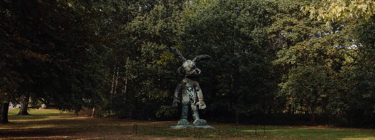 A bronze statue of a figure wearing a large hat and holding a staff stands in a park surrounded by lush green trees. The ground is covered with fallen leaves, indicating the change of seasons. Soft sunlight filters through the foliage.