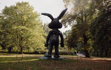A large, whimsical bronze sculpture of a rabbit stands in a park, facing away from the viewer. Surrounded by lush green trees and fallen leaves, the scene captures a serene autumn atmosphere.
