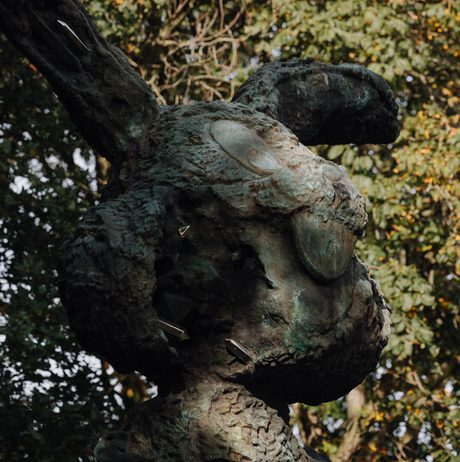 A large, abstract bronze sculpture of a rabbit stands amidst greenery. The figure features textured surfaces and hollow sections, with a whimsical expression. Sunlight filters through the surrounding trees, highlighting the sculpture's details.