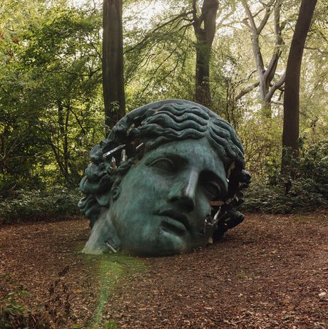 A large, weathered bronze sculpture of a head lies partially buried in a forest clearing. Sunlight filters through the trees, illuminating the serene yet fragmented features of the sculpture, surrounded by lush greenery and fallen leaves.
