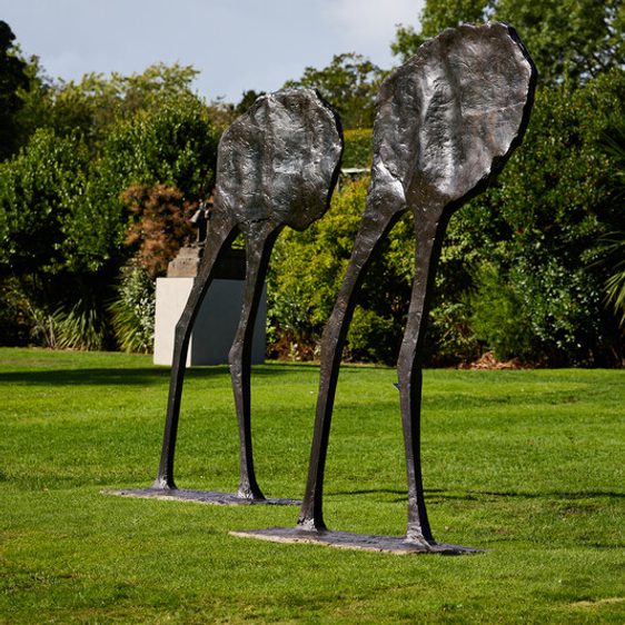Two abstract bronze sculptures resembling elongated figures with leaf-like heads stand on a grassy area. The background features lush greenery and trees under a partly cloudy sky, creating a serene outdoor setting.