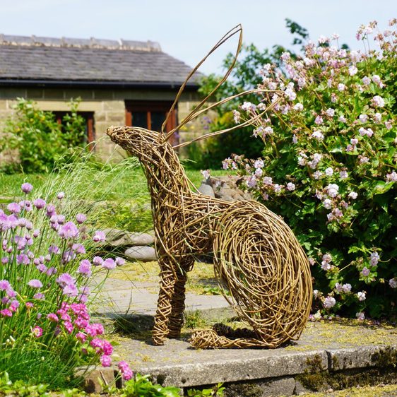 A woven rabbit sculpture sits among colorful flowers in a garden. The rabbit, made of twigs, is positioned on a stone path, with a backdrop of a rustic building and lush greenery, creating a serene outdoor scene.