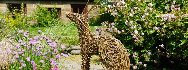 A woven rabbit sculpture sits among colorful flowers in a garden. The rabbit, made of twigs, is positioned on a stone path, with a backdrop of a rustic building and lush greenery, creating a serene outdoor scene.
