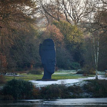 A large, dark sculpture resembling a human head stands in a serene landscape, surrounded by trees and greenery. The ground is lightly dusted with snow, and a calm body of water reflects the tranquil scene.