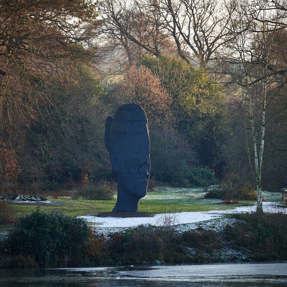A large, dark sculpture resembling a human head stands in a serene landscape, surrounded by trees and greenery. The ground is lightly dusted with snow, and a calm body of water reflects the tranquil scene.