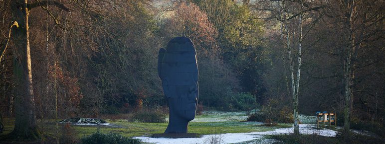 A large, dark sculpture resembling a human head stands in a serene landscape, surrounded by trees and greenery. The ground is lightly dusted with snow, and a calm body of water reflects the tranquil scene.