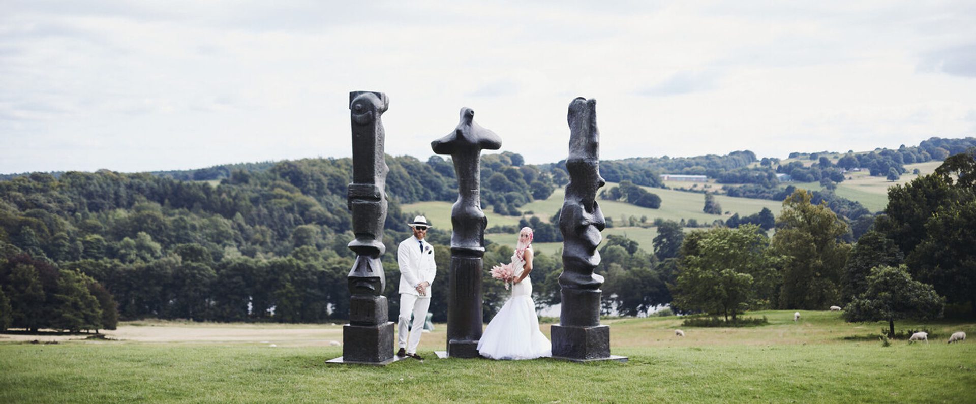 A bride in a white gown and a groom in a white suit stand beside a group of large, abstract sculptures in a green landscape. Rolling hills and sheep are visible in the background under a cloudy sky.