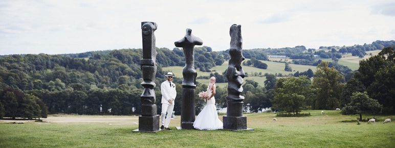 A bride in a white gown and a groom in a white suit stand beside a group of large, abstract sculptures in a green landscape. Rolling hills and sheep are visible in the background under a cloudy sky.