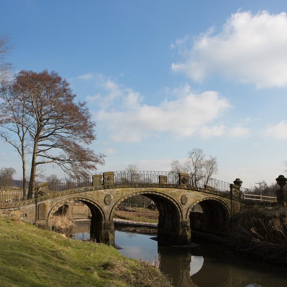 A stone bridge arches over a calm river, surrounded by bare trees and grassy banks. The sky is bright with fluffy clouds, creating a serene landscape. The bridge features decorative stonework and railings, adding to its charm.