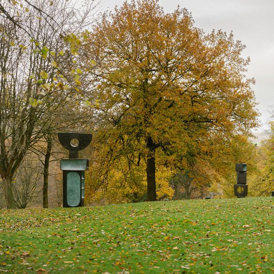 A serene park scene featuring several abstract sculptures set against a backdrop of autumn trees with vibrant orange and yellow leaves. The sculptures vary in shape and size, creating a harmonious blend of art and nature.