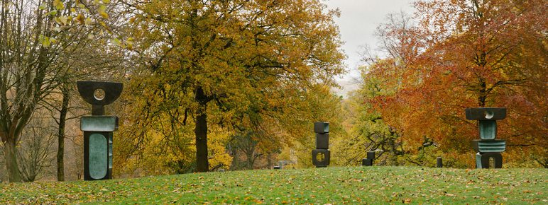 A serene park scene featuring several abstract sculptures set against a backdrop of autumn trees with vibrant orange and yellow leaves. The sculptures vary in shape and size, creating a harmonious blend of art and nature.