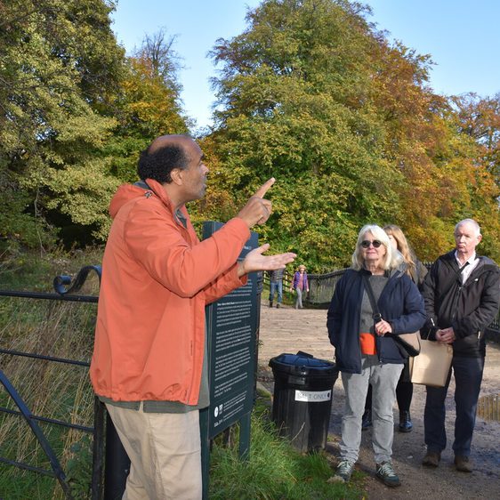 A diverse group of people listens attentively to a speaker in an orange jacket, gesturing animatedly. The scene is set in a park with vibrant autumn foliage in the background, under a clear blue sky.