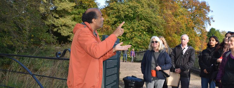 A diverse group of people listens attentively to a speaker in an orange jacket, gesturing animatedly. The scene is set in a park with vibrant autumn foliage in the background, under a clear blue sky.