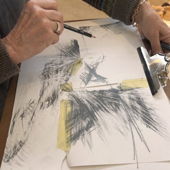 A close-up of a person's hand holding a pencil, sketching on a sheet of paper. The paper features abstract charcoal drawings, with some areas taped down. The individual wears a knitted sweater with colourful stripes.