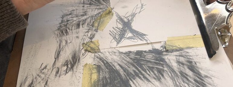 A close-up of a person's hand holding a pencil, sketching on a sheet of paper. The paper features abstract charcoal drawings, with some areas taped down. The individual wears a knitted sweater with colourful stripes.