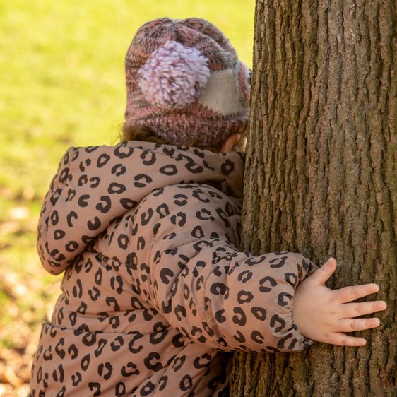 A young child wearing a leopard-print coat and a knitted hat with pom-poms is playfully hugging a tree. The scene is set in a park with green grass and fallen leaves, capturing a moment of joy in nature.