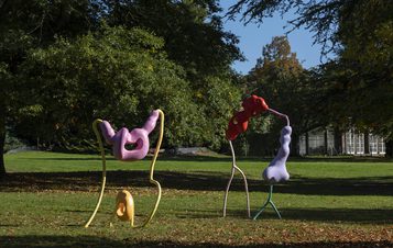 Three abstract sculptures in vibrant colours—pink, red, and purple—stand on a grassy area surrounded by trees. The playful shapes feature curved lines and organic forms, contrasting with the natural setting and a building in the background.