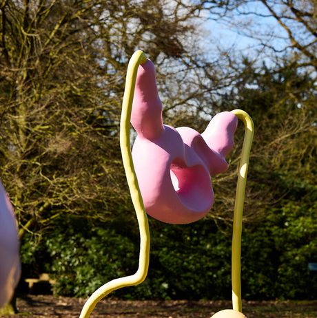 A whimsical sculpture in pastel pink and yellow, resembling abstract forms, stands against a backdrop of trees. The artwork features smooth curves and playful shapes, creating a vibrant contrast with the natural surroundings.