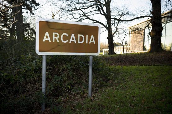 A brown sign reading "ARCADIA" stands on a grassy area, surrounded by shrubs. In the background, bare trees and a modern building are visible, with a clear sky above, indicating a tranquil outdoor setting.