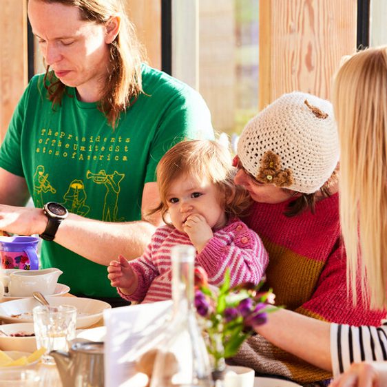 A family gathering at a wooden table, featuring a man in a green t-shirt, a woman in a pink and red striped sweater holding a toddler. The toddler is eating, while others enjoy a meal, surrounded by plates and glasses. Natural light fills the space.