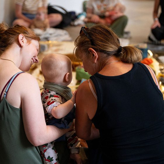 Two women sit closely together, one holding a baby, as they engage in a communal activity. The setting is warm and inviting, with soft lighting and a variety of textures around them. Other participants are visible in the background, creating a sense of community.