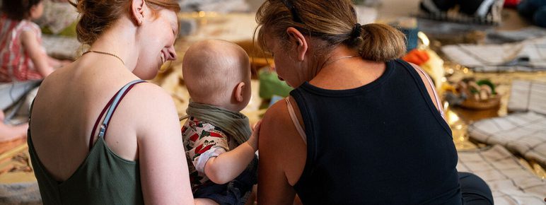 Two women sit closely together, one holding a baby, as they engage in a communal activity. The setting is warm and inviting, with soft lighting and a variety of textures around them. Other participants are visible in the background, creating a sense of community.