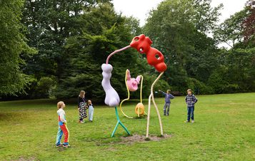 A vibrant outdoor sculpture installation features abstract, colourful forms resembling playful figures. Children and adults interact with the artwork in a lush green park, surrounded by trees and a serene atmosphere.