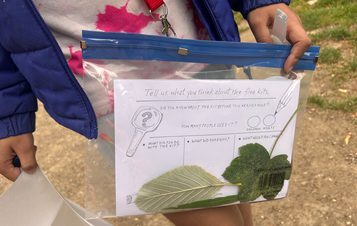 A person holds a clear plastic bag containing a questionnaire about free kits, featuring a magnifying glass illustration and two leaves. The sheet asks for feedback on the kit's usage and encourages sharing thoughts.