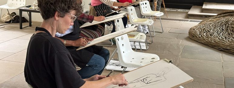 A group of artists sits in a spacious room, each focused on drawing from a model. They use easels and sketch pads, with a variety of styles and techniques visible. The setting features large windows and minimalist decor, creating a creative atmosphere.